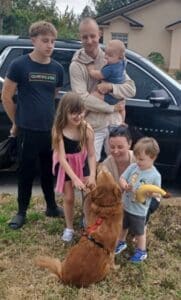 An adult golden retriever is sitting outside with its back facing the camera, and looking up at its new family. A mom is squatting and petting the dog, with an excited child on either side. A smiling dad is standing behind them, holding a baby, with a teenager to his side. Smiles all around.