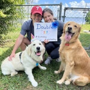 Adoptive parents sitting with two goldens. The parents are holding a sign for a happy birthday and gotcha day. One of the goldens is wearing a party hat.