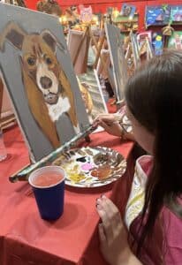 A woman painting a portrait of a brown and white dog, with a paint palette and water cup on a red table.