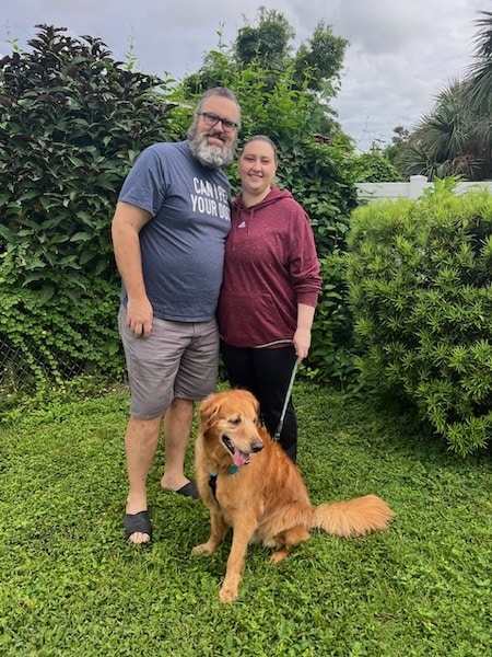 A man and a woman standing outside while holding the leash of a happy golden retriever sitting in front of them.