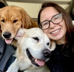 Smiling woman poses with two happy Golden Retrievers with their tongues out, beside her inside a car.