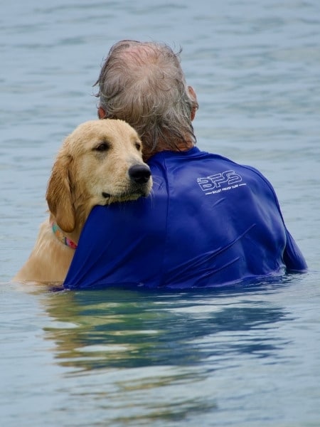 Golden retriever resting its head on a man’s shoulder while both stand in calm ocean water.
