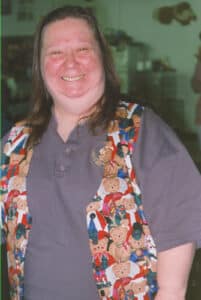 A smiling woman indoors wearing a patterned vest over a gray shirt.