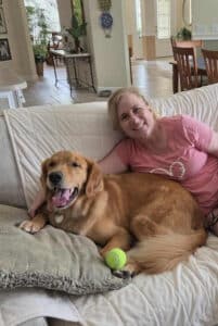 Golden is laying on a white couch, resting on a pillow with a yellow tennis ball close. One of his human friends is also sitting on the couch next to him hugging him.