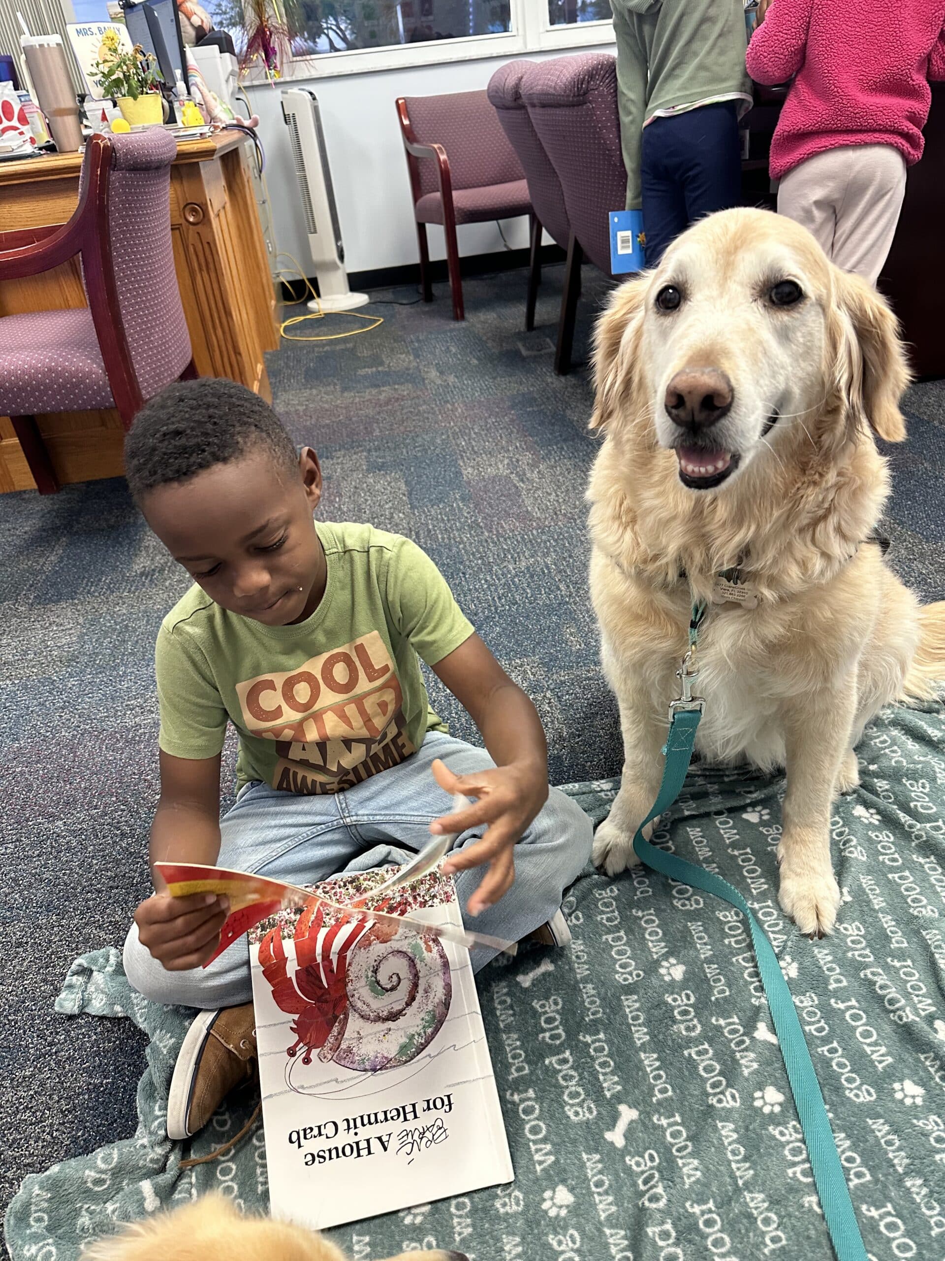 Golden retriever wearing a Hearts of Gold vest sitting next to a boy reading a book in a classroom.