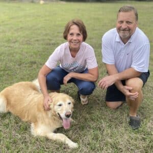 Golden lying on grass with two people. Golden retriever resting on grass between two adults kneeling beside him, tongue out and relaxed.