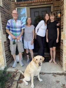 Family of four standing in front of a house with a tan dog on a leash, all smiling at the camera