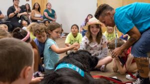 Children sitting together watching a Cavachon wearing a Hearts of Gold therapy vest during a therapy visit, part of the Hearts of Gold therapy team.
