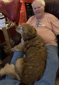 Golden retriever with a red coat stretched across a person’s lap in a recliner, wearing a black collar with a red tag, relaxed and content while the person in an orange shirt and jeans rests with them.