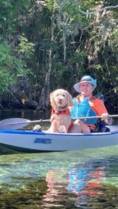 Goldendoodle riding in a kayak with a handler on a clear river, part of the Hearts of Gold therapy team.
