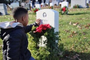 A child kneels to place a wreath with a red bow at a veteran’s gravestone.