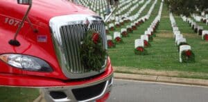 A red truck displays a wreath on its grille while rows of white headstones, each adorned with wreaths, stretch across the cemetery behind it.