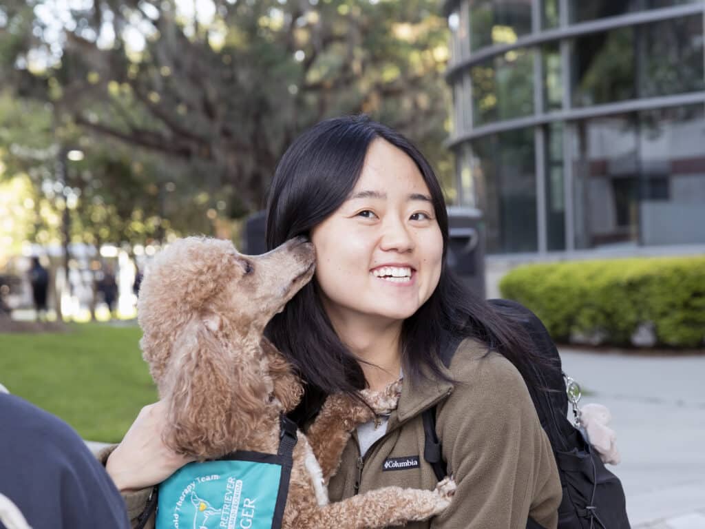 A smiling student holding a poodle named Ginger wearing a Hearts of Gold vest that is nuzzling her cheek.