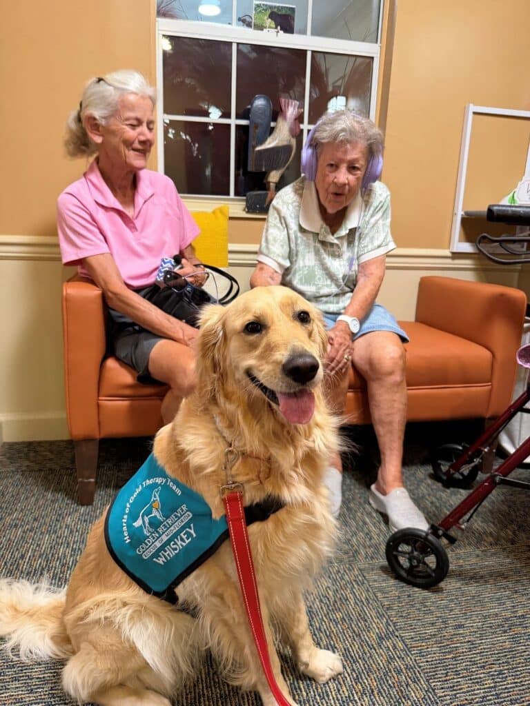 Whiskey, a Golden Retriever blend therapy dog, sits smiling in front of two women during a senior living visit.