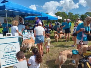 Outdoor community event with volunteers, visitors, and golden retriever therapy dogs gathered around blue canopy tents.