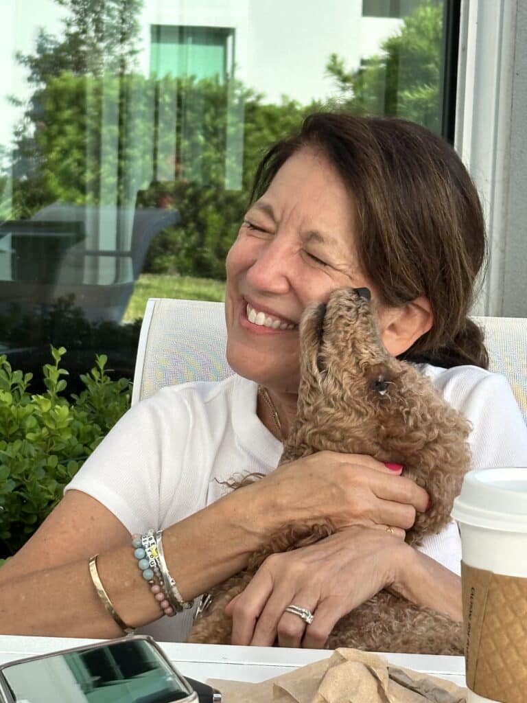 Woman smiling while holding and embracing a curly-coated brown poodle therapy dog.